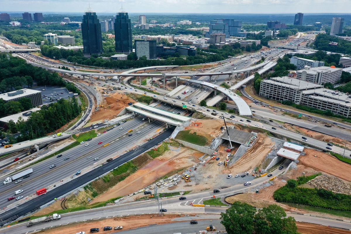 Aerial view of a busy highway interchange under construction near tall city buildings, daytime with greenery